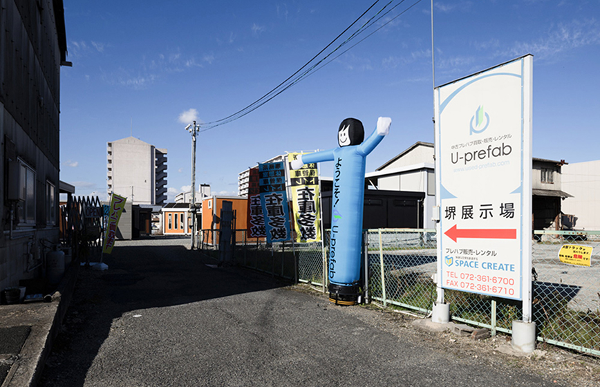 大阪堺展示場の様子 ようこそ！と書かれた人型のバルーンやのぼりがあり、奥に簡易住宅の展示が連なっている